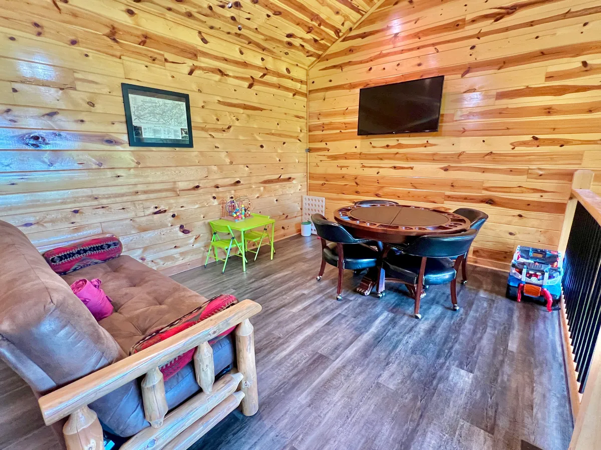 This interior shot focuses on the loft's poker table, surrounded by a comfy sofa and a dedicated children's table. The room's wooden walls and large windows keep the scene bright and welcoming. Family members are seen playing cards, relaxing together, or watching TV, exemplifying the room’s versatility for both group games and quiet movie nights.