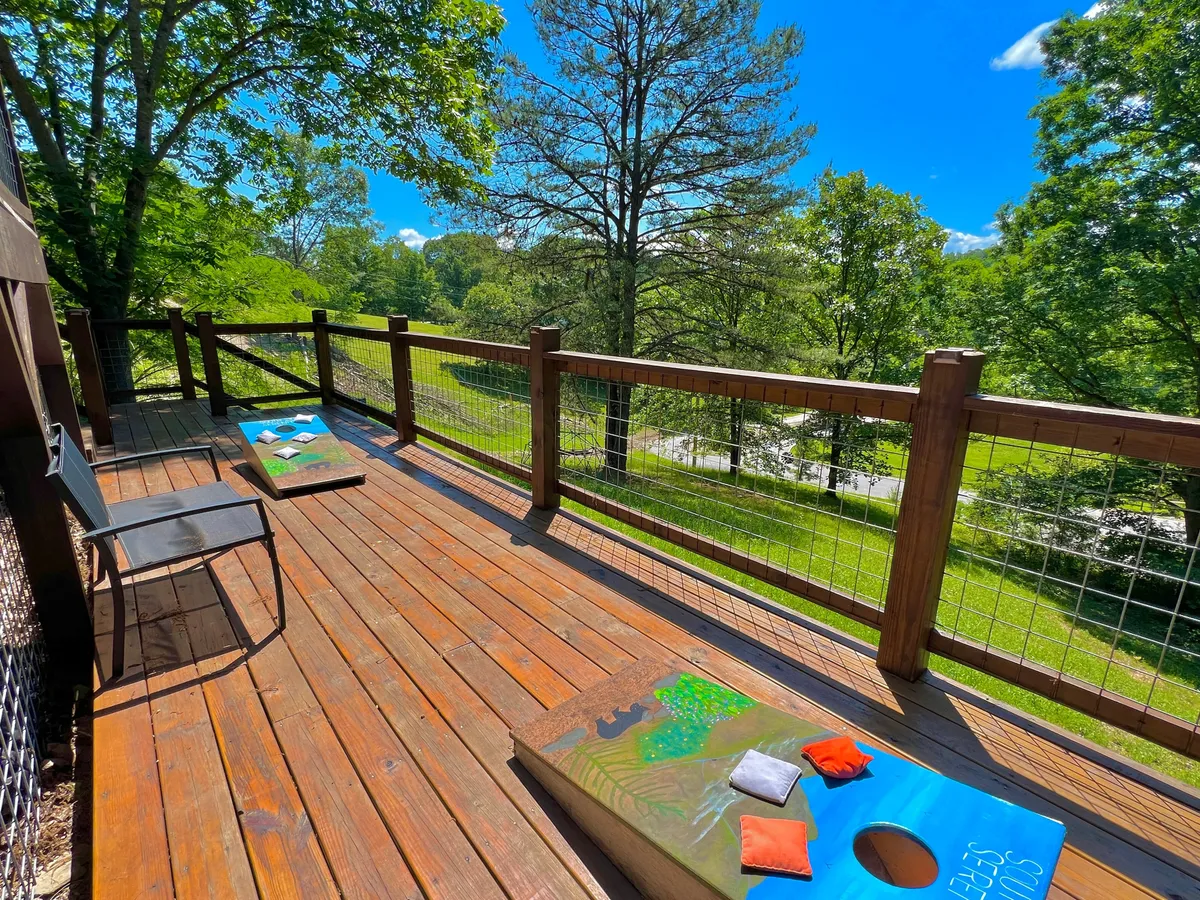 A wooden deck features cornhole boards set up for play, surrounded by lush trees and outdoor seating. This space is perfectly designed for friendly competitions and relaxed gatherings with friends, with nature providing a scenic backdrop.