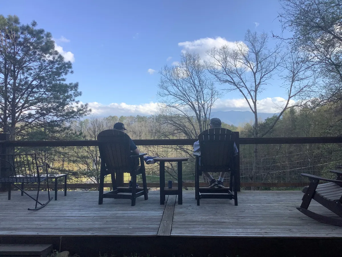 This image shows two people sitting in Adirondack chairs on a spacious wooden deck, overlooking a breathtaking expanse of distant mountains and tree-lined valleys. The natural setting is peaceful and inviting, with the deck positioned perfectly for taking in sunsets or stargazing beneath open skies.