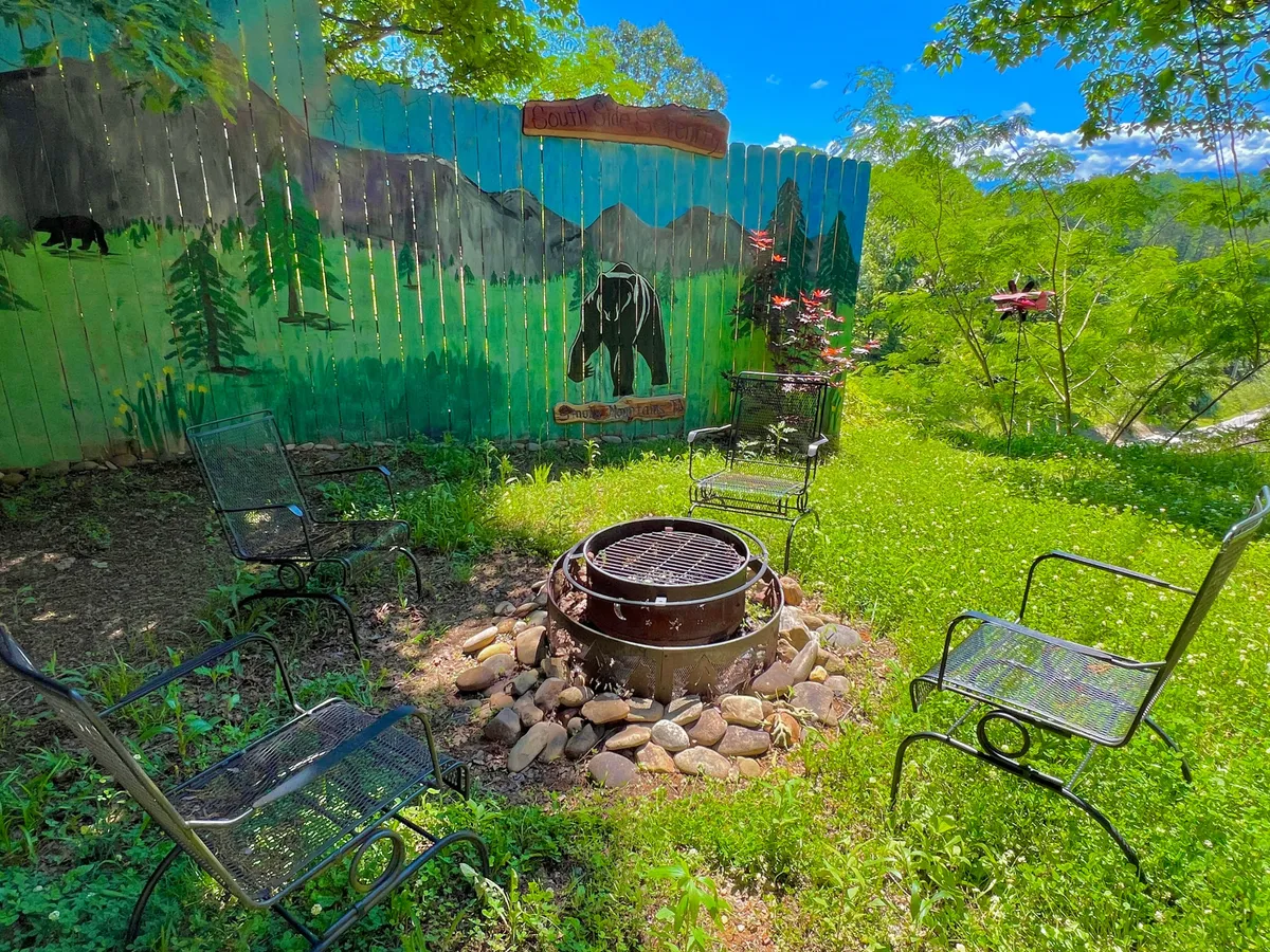 A cozy outdoor area with a fire pit is surrounded by comfortable seating and lush greenery, featuring a decorative mural as a vibrant backdrop. The arrangement invites groups to gather for storytelling, marshmallow roasting, and enjoying nature together.