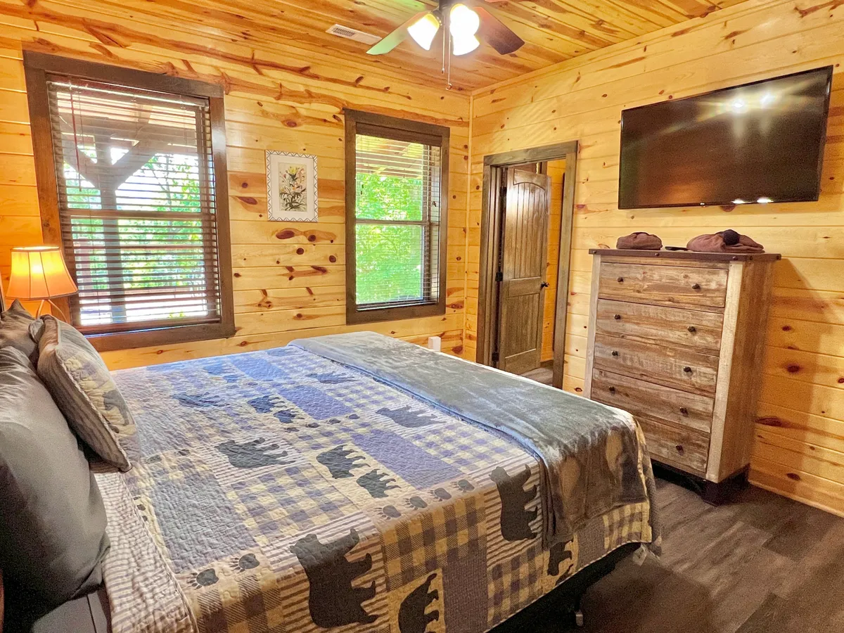 A cozy bedroom featuring a bed with a patterned blanket, a wooden dresser, windows with blinds letting in natural light, and a TV mounted on the wall. The room feels inviting and comfortable, ideal for relaxation or enjoying a movie night in bed.