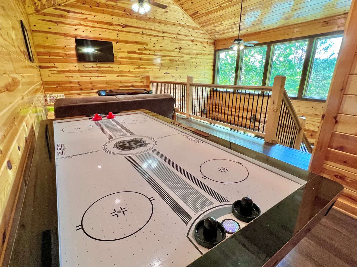 This image captures the main area of the loft game room featuring a prominent air hockey table set against rustic wooden walls and bathed in natural light from large windows. Individuals are engaged in playing air hockey, while others socialize and enjoy the relaxed ambiance. The background hints at a big-screen TV, contributing to the entertainment atmosphere.