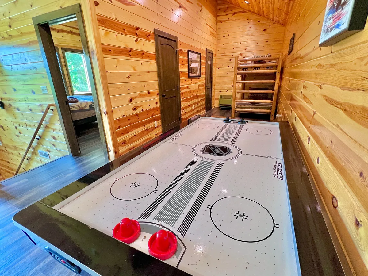 A lively game room features a central air hockey table surrounded by rustic wooden walls and doorways leading to other rooms. The environment feels energetic and inviting, hinting at friendly competition and family tournaments.