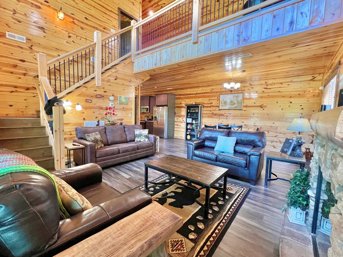 A comfortable living area with leather sofas arranged around a stone fireplace, a central coffee table, a wooden staircase, and warm lighting. The atmosphere is welcoming and ideal for family gatherings or unwinding after travels.
