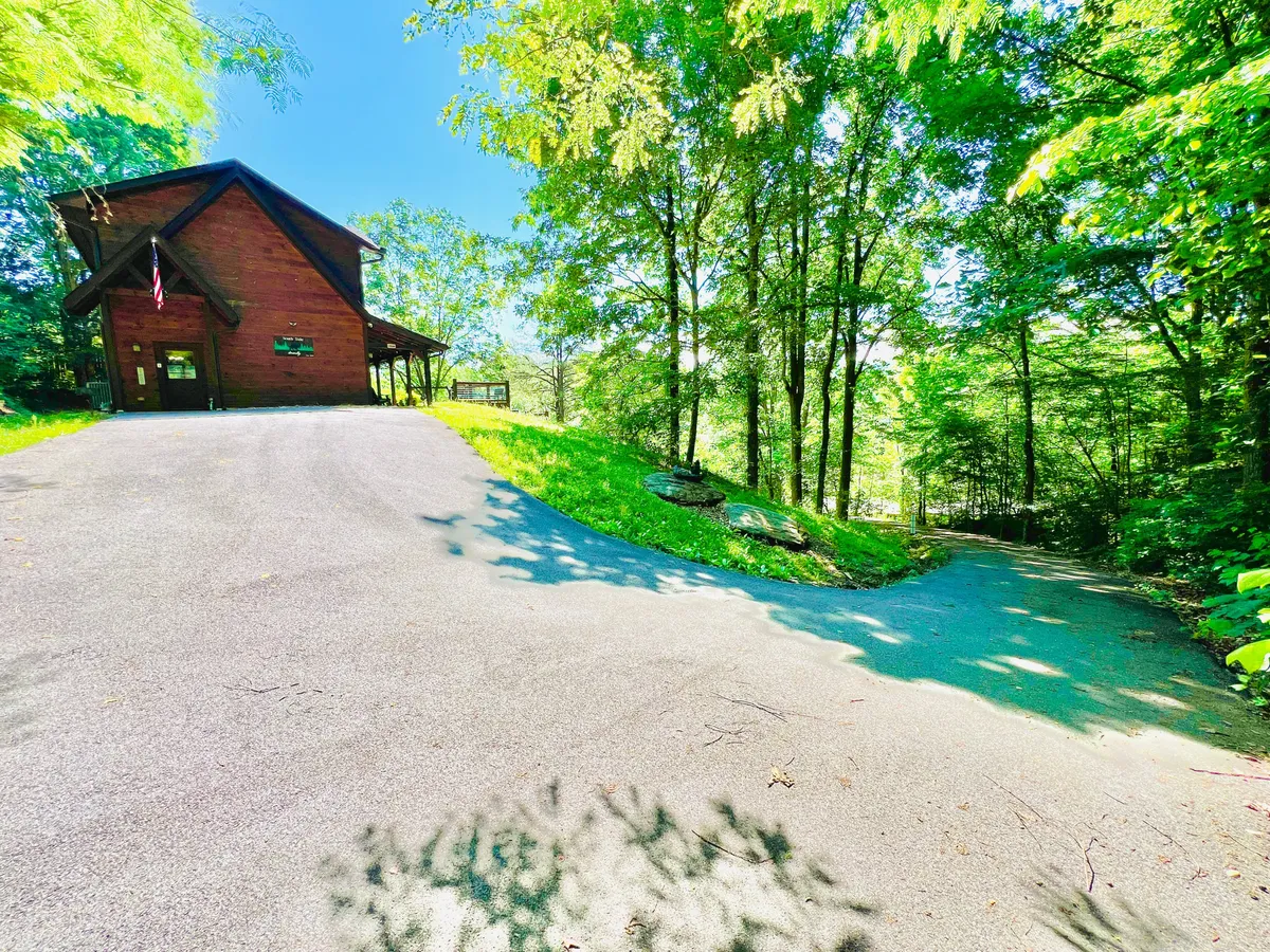 The image highlights the exterior of a charming wooden cabin set among trees, with a paved driveway offering easy parking. The peaceful yard is an inviting spot for group arrivals, outdoor gatherings, or relaxing in the fresh mountain air.