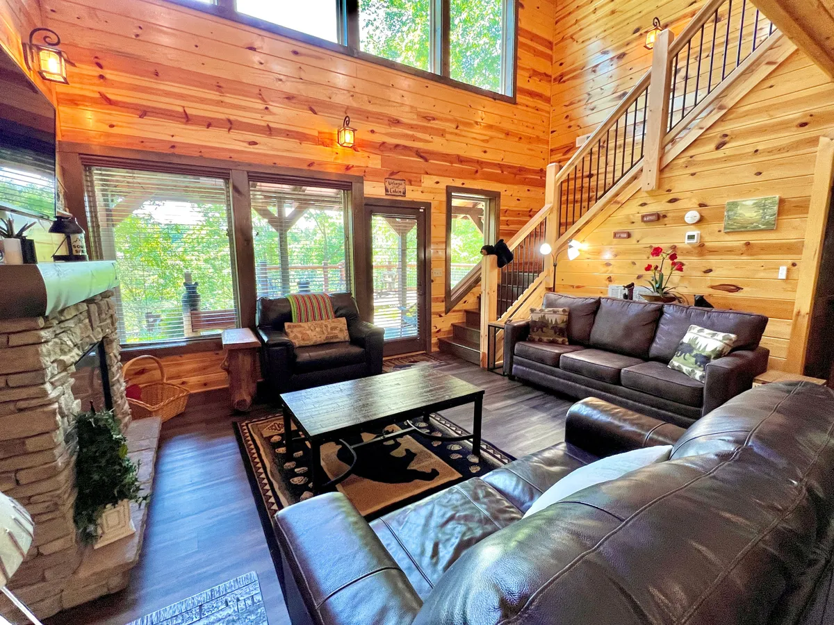 A spacious, welcoming living space featuring a stone fireplace, large windows offering views of the outdoors, a wooden staircase, cozy furniture, and a rug with a bear motif. The setup is perfect for relaxing moments or lively gatherings.