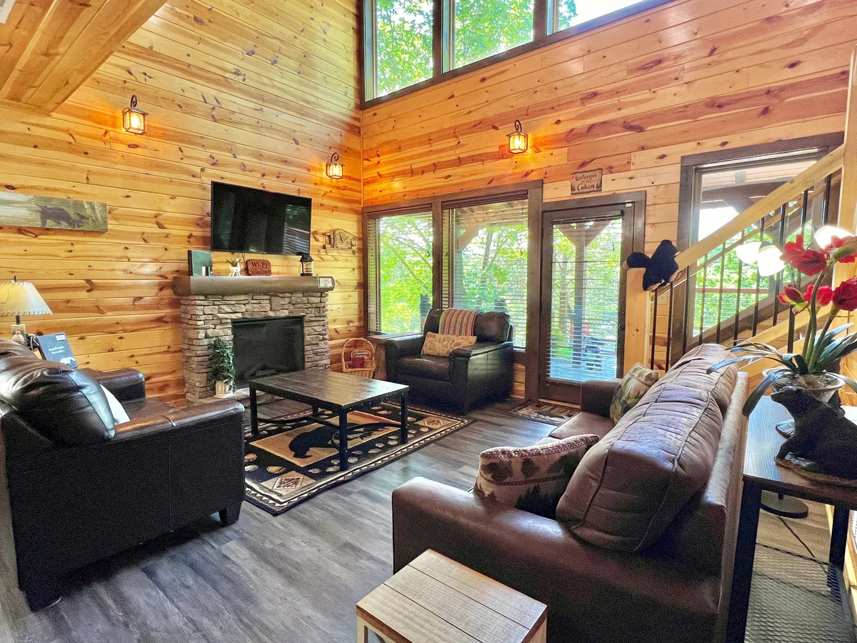 A warm and inviting living room featuring a stone fireplace, large windows letting in natural light, comfortable seating, and a decorative rug. The space is arranged to encourage conversation and relaxation, with the fireplace as a focal point and ample room for gathering together.