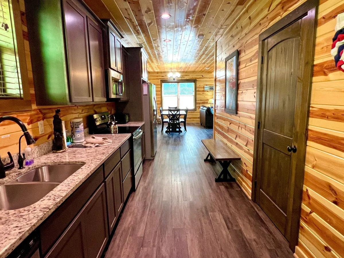 This shot captures an open kitchen and dining area, featuring a granite countertop, dark cabinetry, dining area with a wooden bench, and large windows that allow sunlight to fill the space. The setup is clearly designed for cooking, dining, and socializing in a cozy environment.