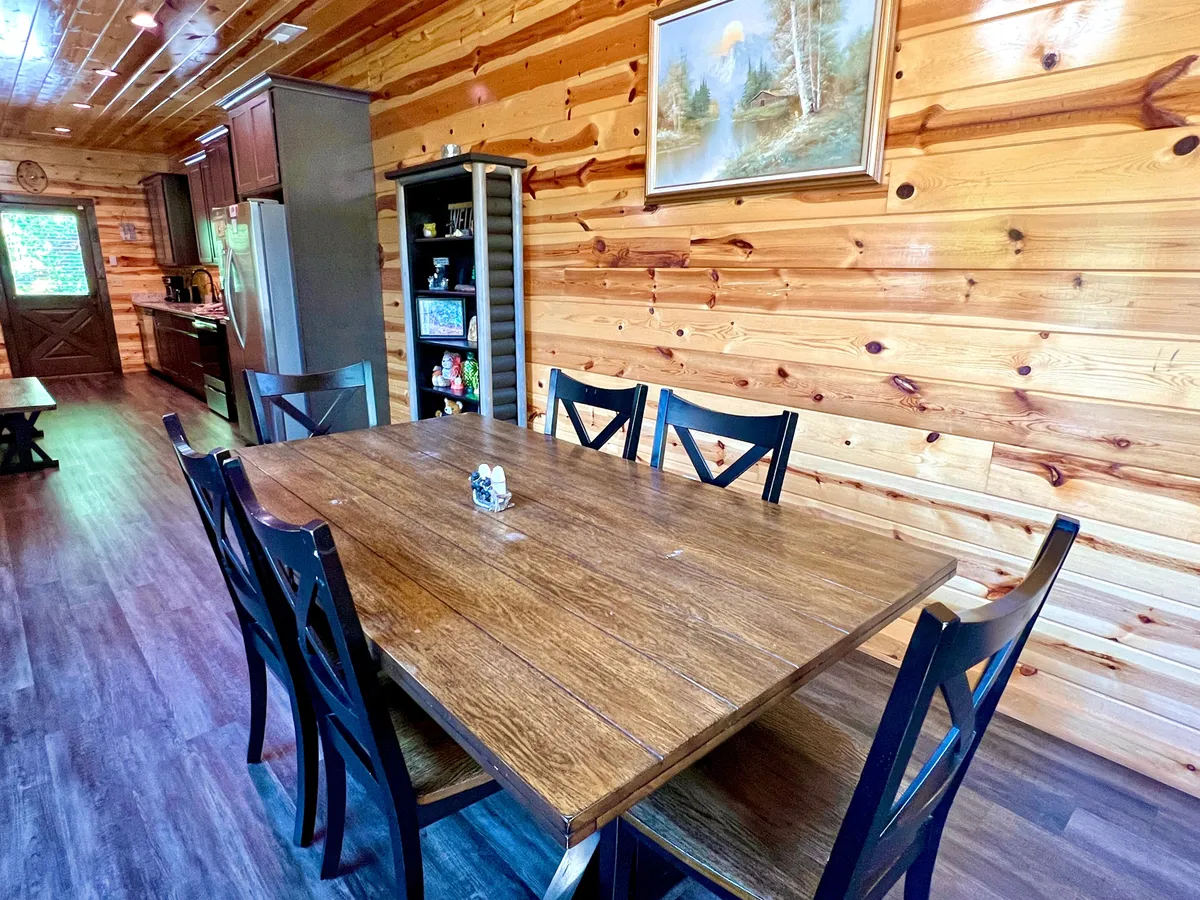 A cozy dining area with a large wooden table, matching chairs, and a visible kitchen in the background. Warm lighting and wall art add to the inviting feel, making it the perfect spot for family meals and casual conversations.